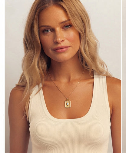 A blonde woman wearing a cream ribbed tank top and a delicate gold necklace with a rectangular pendant, photographed against a light neutral background.
