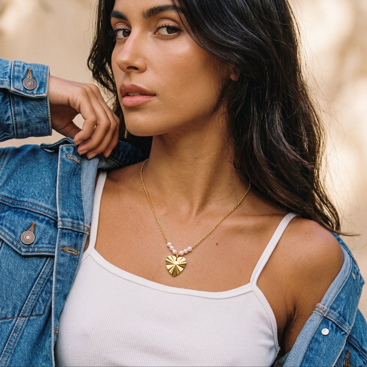 A woman wearing a delicate gold necklace with a flower-shaped pendant and small pink beads, styled with a white ribbed tank top and an open blue denim jacket.