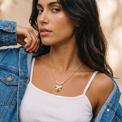 A woman wearing a delicate gold necklace with a flower-shaped pendant and small pink beads, styled with a white ribbed tank top and an open blue denim jacket.