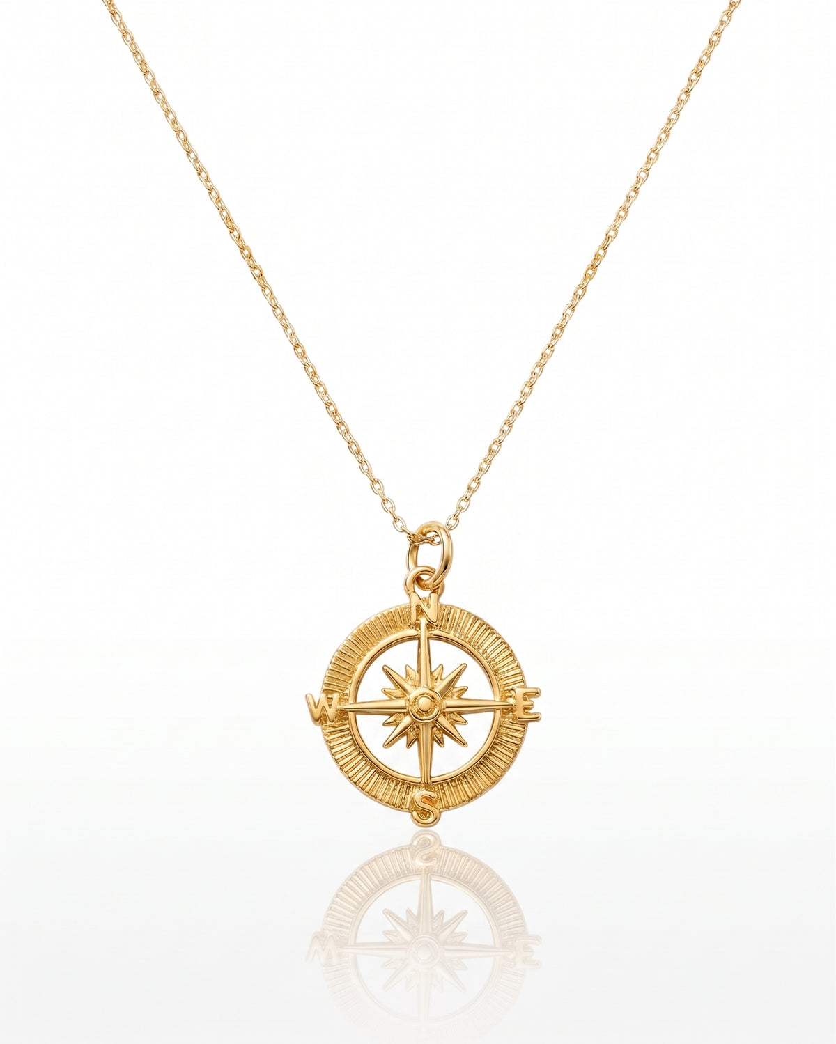 A gold compass pendant necklace on a delicate gold chain, displayed against a clean white background with a soft reflection below.