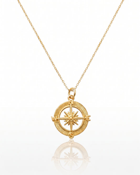 A gold compass pendant necklace on a delicate gold chain, displayed against a clean white background with a soft reflection below.