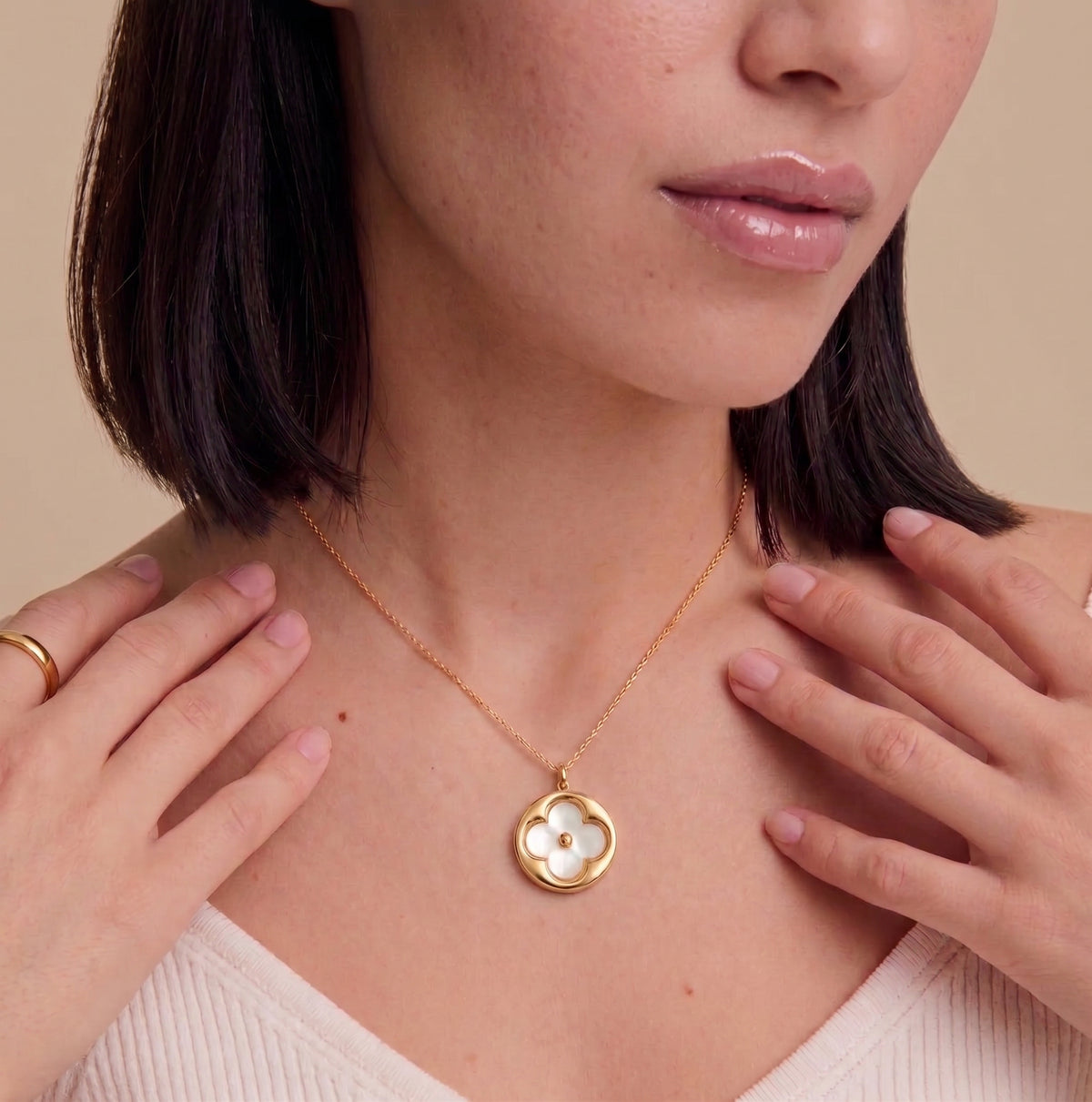 A close-up of a woman wearing a delicate gold necklace with a flower-shaped pendant featuring a white inlay, set against a warm beige background.