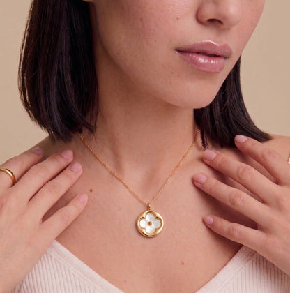 A close-up of a woman wearing a delicate gold necklace with a flower-shaped pendant featuring a white inlay, set against a warm beige background.