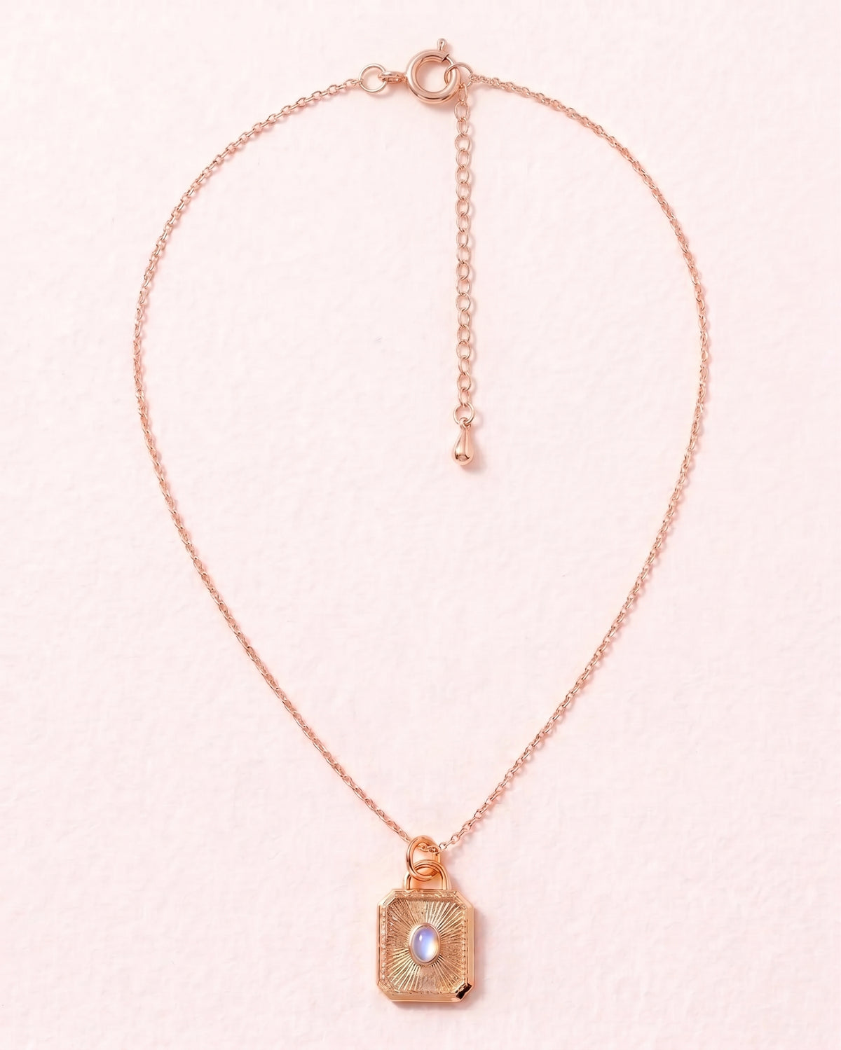 A delicate rose gold chain necklace featuring a small square pendant with a textured gold-toned frame and a central iridescent pearl, displayed on a soft pink background.