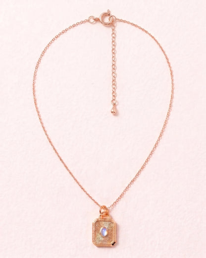 A delicate rose gold chain necklace featuring a small square pendant with a textured gold-toned frame and a central iridescent pearl, displayed on a soft pink background.