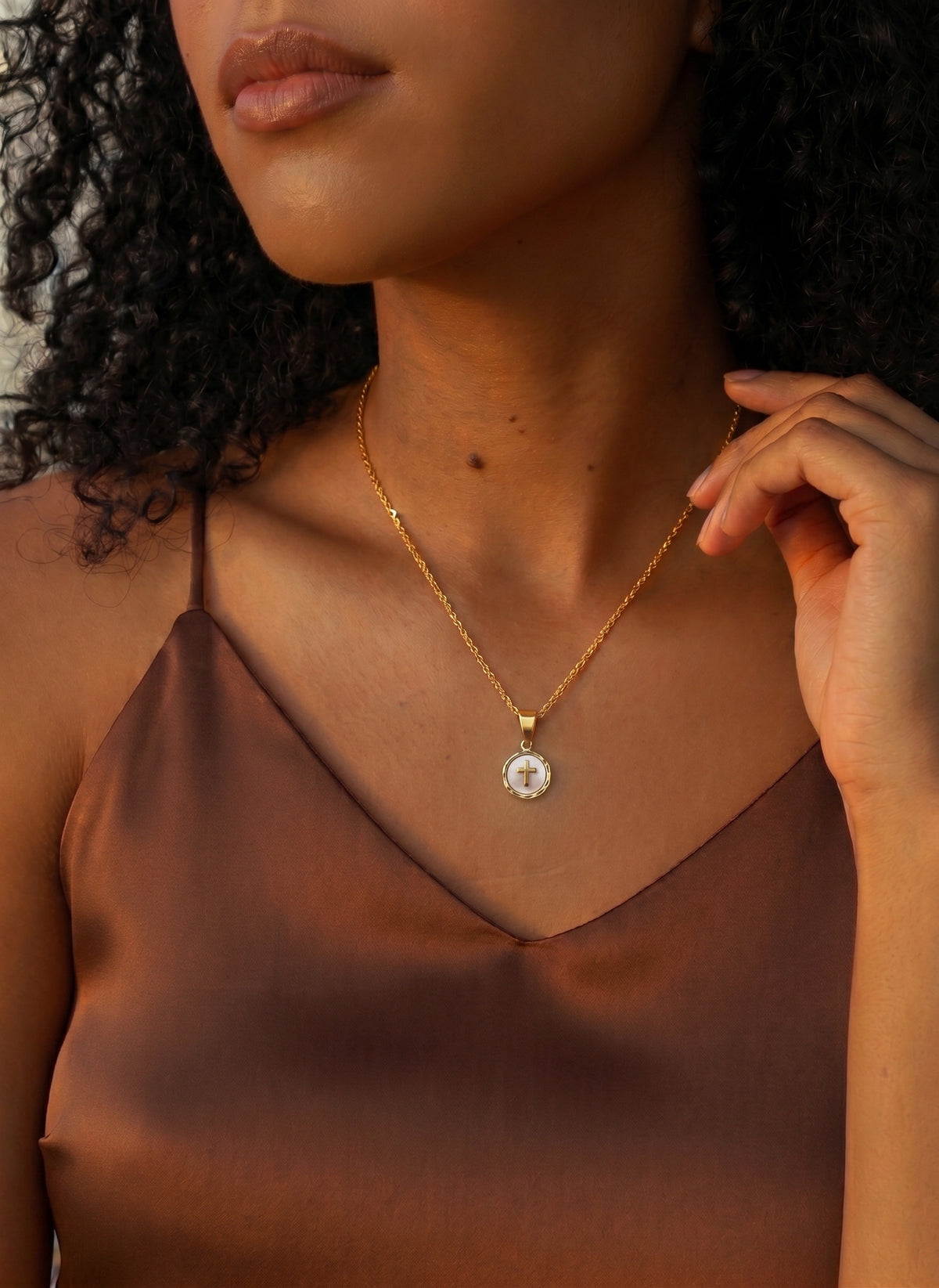 A close-up of a woman wearing a delicate gold chain necklace with a round white pendant featuring a central gold cross, styled with a brown satin camisole.