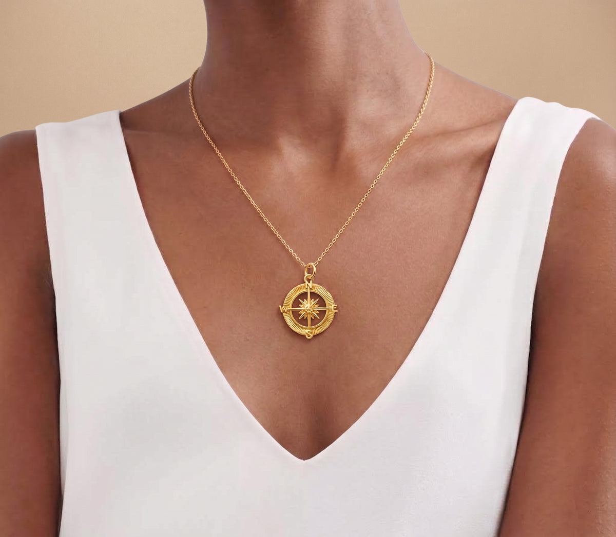 A gold compass pendant necklace worn by a model in a white V-neck top, shown against a warm beige background.