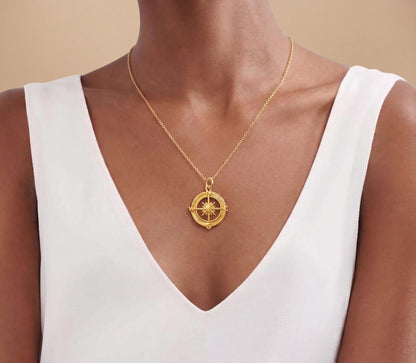A gold compass pendant necklace worn by a model in a white V-neck top, shown against a warm beige background.