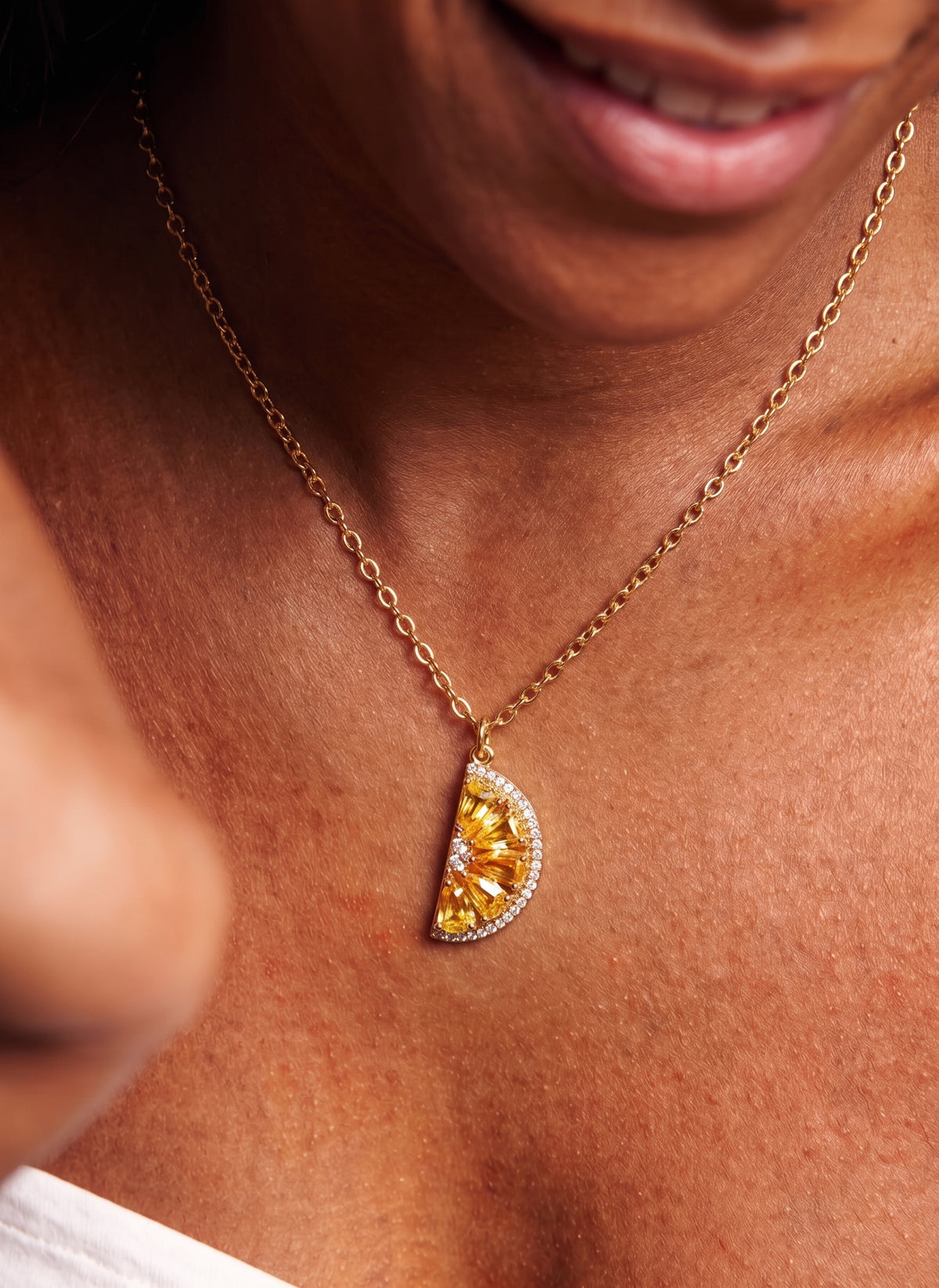 A close-up lifestyle shot of a model wearing a gold chain necklace with a semi-circular pendant featuring yellow gemstones and a halo of small clear stones.