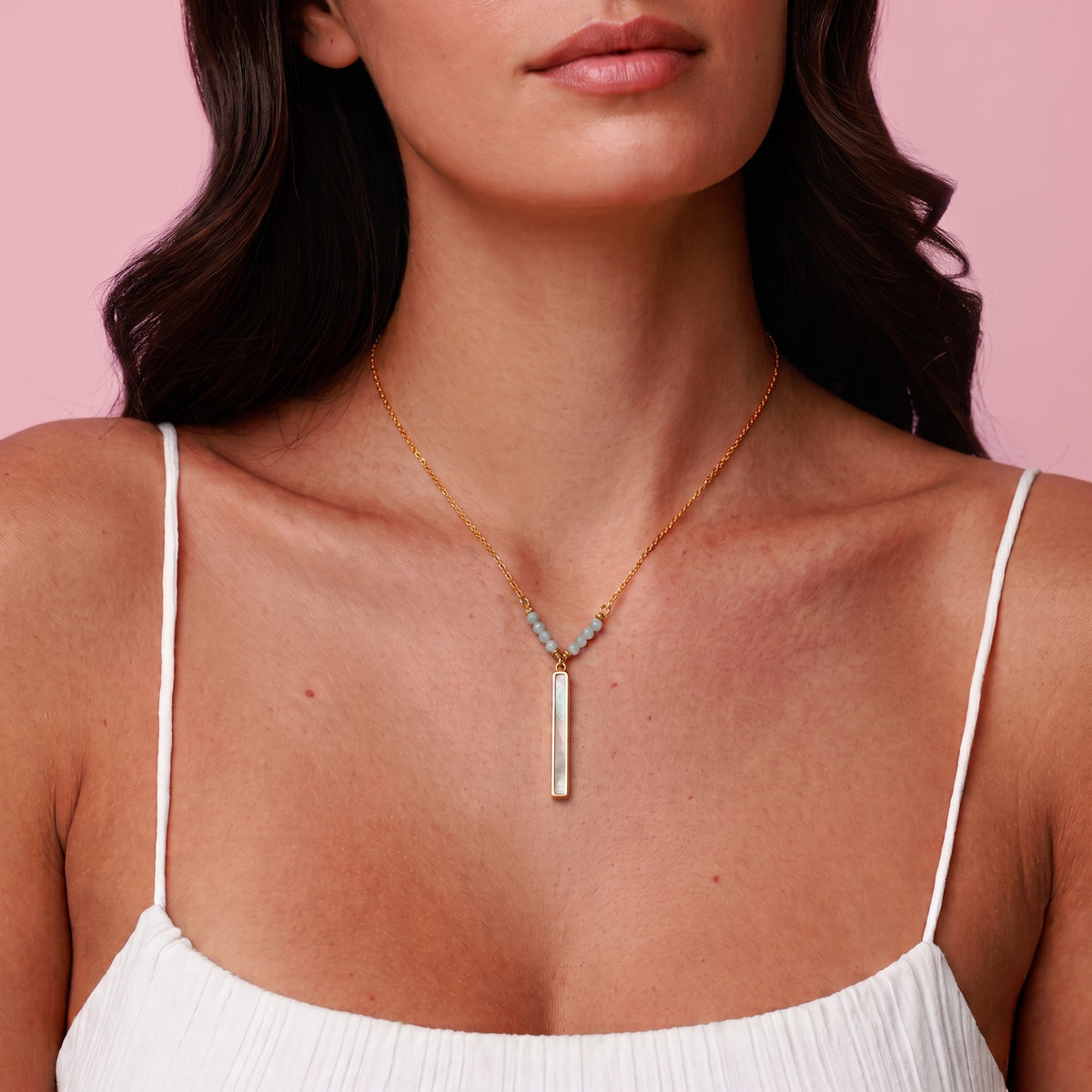 A close-up of a model wearing a delicate gold necklace with a vertical bar pendant and small light blue stones at the V-shaped top, paired with a white spaghetti-strap top against a solid pink background.