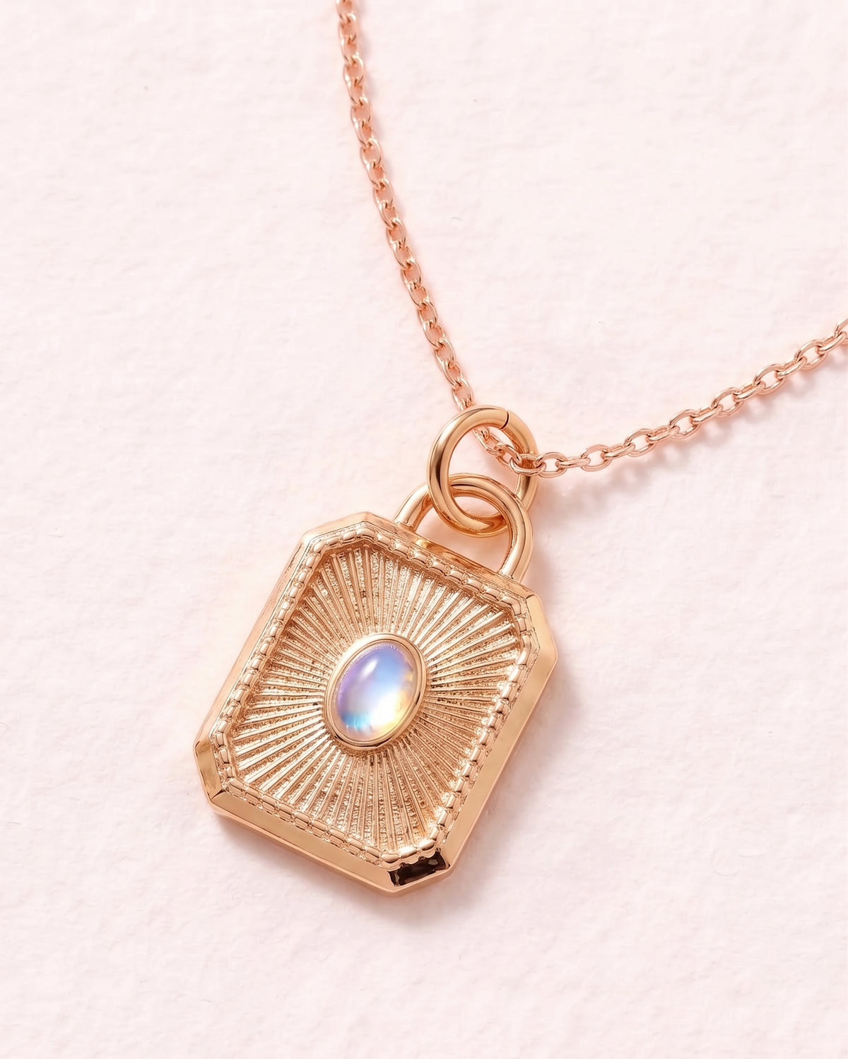 A rose gold pendant necklace featuring a textured, geometric shield-shaped charm with a central iridescent opal-like stone, shown on a delicate chain against a light pink background.